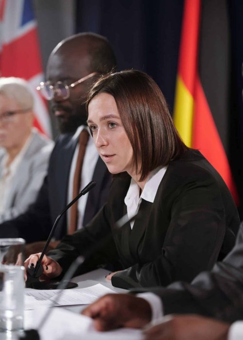 Young female politician in formalwear speaking in microphone while sitting among intercultural foreign colleagues at conference or summit